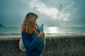 Mother with baby in sling looking over wall by the sea