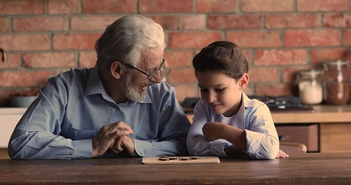 Happy elderly mature 60 hoary man in eyeglasses playing checkers draughts on wooden board with joyful little grandson, enjoying creative game on weekend leisure time, having fun together at home.