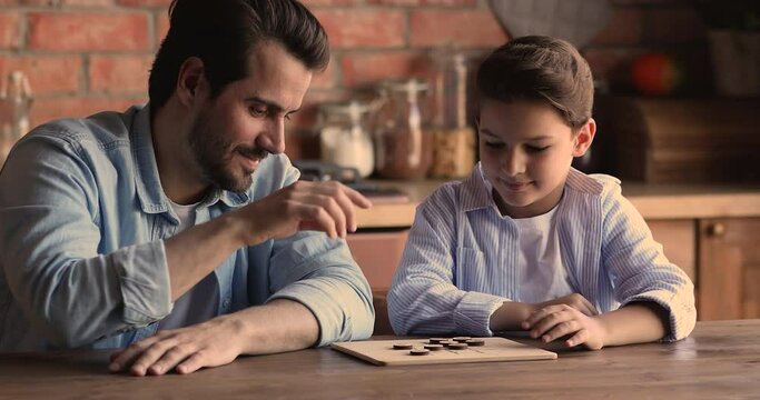 Joyful young father playing checkers draughts on wooden board with smart concentrated little school aged son, enjoying family playtime domestic activity, entertaining improving logic skills at home.