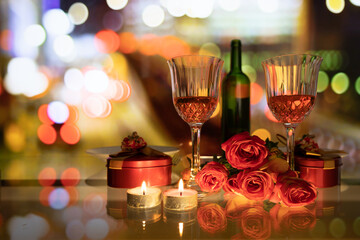 red roses, candles in table setting with wine glass and bottle. blurred colorful city night in background.