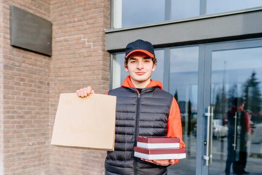 Delivery, mail and people concept. Young male worker, delivery man carrying brown craft paper bag for takeaway and boxes with sushi to customer in house on the doorstep, outdoor.