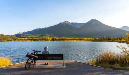 Tourist resting on wooden bench, relaxing at Vermilion Lakes lakeshore in autumn foliage season sunset time. Cycling in Banff National Park Legacy Trail. Canadian Rockies, Alberta, Canada.