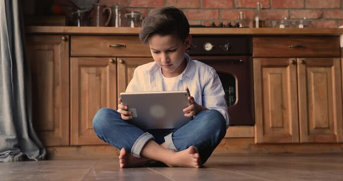 Happy little cute kid boy sitting barefoot on warm heated wooden floor, involved in playing online video game on digital computer touchpad, enjoying communicating in social network, tech addiction.