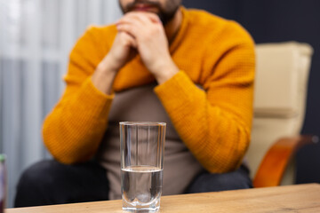 Glass of water on the background of a pensive caucasian man holding his hands under his beard. Indoor shot. Selective focus