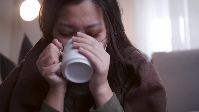 Sick Asian woman drinking lukewarm water in times of suffering has chilling black eyelids all over her body due to a cold and uses masks and blankets to warm and quarantine herself