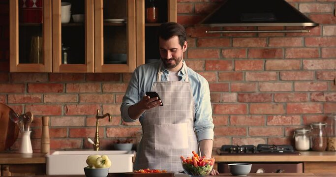 Distracted from cooking smiling young handsome man in apron involved in communicating in social network using mobile phone or web surfing interesting recipe for dinner, preparing food alone at home.