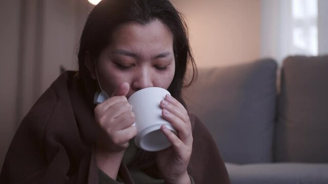 Sick Asian woman drinking lukewarm water in times of suffering has chilling black eyelids all over her body due to a cold and uses masks and blankets to warm and quarantine herself