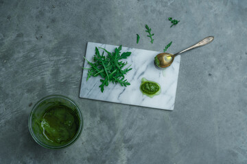 
Wild garlic sauce on a plate with spoon and organic, vitamin-rich and crunchy ruccola salad on a white marbled background with gray