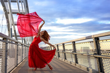 Beautiful redhead female dancer in red long skirt gacefully dance with red silk scarf in her hand on the bridge footpath at dusk