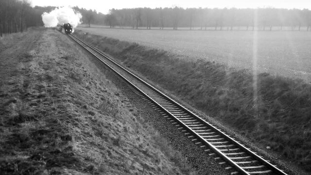 Vintage steam locomotive with old wagons on trail. Black and white footage of train with white smoke on railroad. Retro passenger transport and travel.