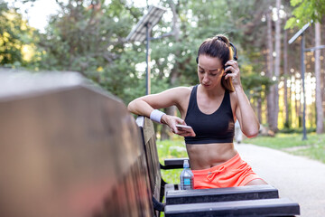 Young strong fitness woman in sportswear resting on bench and listening music on smartphone in park