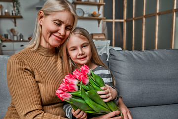 I love you grandma! Grandmother and granddaughter sitting embracing on the couch holding pink tulips. Spring, celebration, mother and woman's day concept