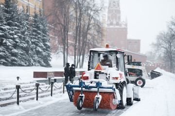 A small red tractor with a brush sweeps snow from the sidewalk near the Kremlin walls during a heavy snowfall. Snowflakes are flying in the air.