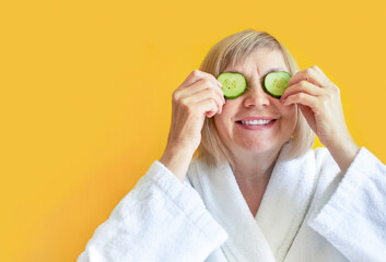 Cheerful senior woman with cucumber slices covering her eyes in front of her eyes on a yellow...