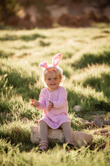 Baby girl wears rabbit ears, sitting in grass.