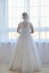 Bride in a apartment in a wedding dress. Bride looks out of the window, wedding day.
Charming bride stand in front of window in a white wedding dress.