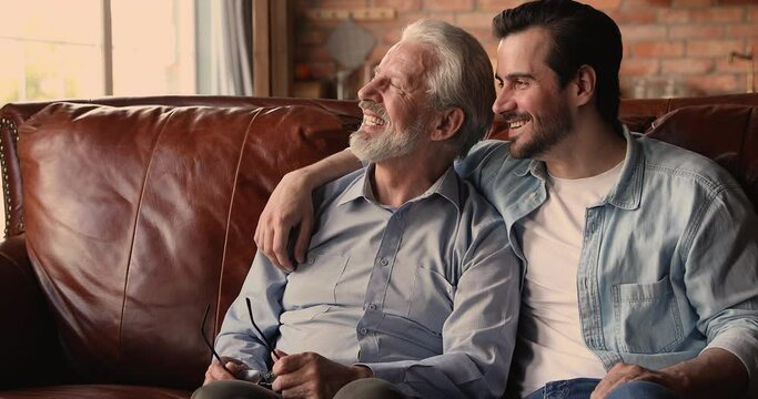 Dreamy bonding sincere middle aged elderly man and affectionate young grownup son looking in distance, discussing life news, enjoying carefree pleasant conversation, resting on cozy sofa at home.