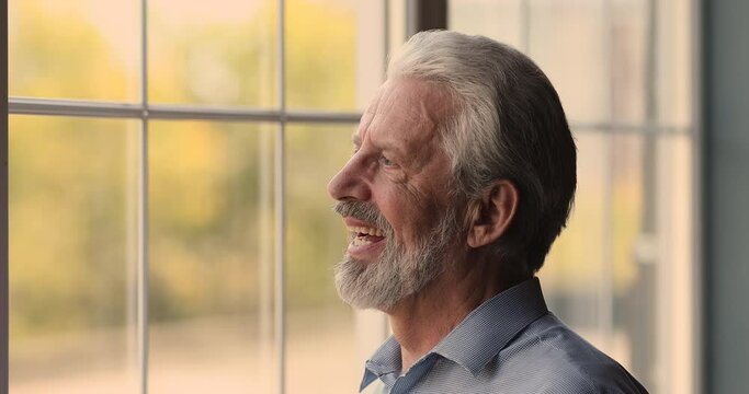 Close up head shot side view dreamy middle aged senior 60s hoary man looking out of window, visualizing future, feeling happy recollecting good memories or waiting for grownup children visit.
