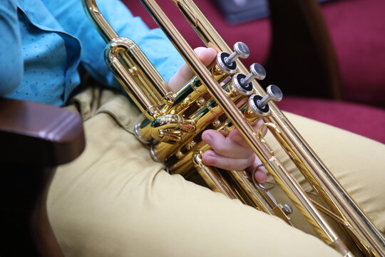 Close-up of a brass musical instrument a shiny gold-colored trumpet between the legs of a man sitting in a chair hand with gentle teenage fingers on the buttons. The musician is resting