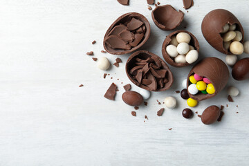 Broken chocolate eggs with candies on white wooden table, flat lay. Space for text