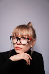 Close-up portrait of a young attractive woman in glasses wearing black sweater. A girl with blonde hair on gray background looking straight in camera