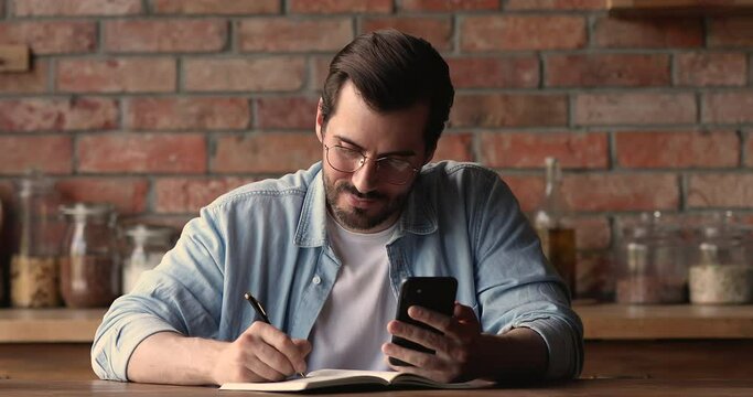 Focused young 30s caucasian man in eyeglasses sitting at table, web surfing information online on smartphone, writing notes in copybook, studying distantly or planning workday alone indoors.