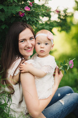 Mother and child daughter in spring garden. Young woman and baby girl together standing near the blooming bush with pink rose flowers. Baby touching flowers on bush. summer time