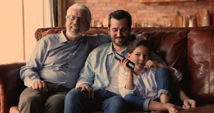 Happy multigenerational male family resting on cozy sofa, watching funny comedian movie together in living room. Smiling carefree young man spending free weekend time with old father and small son.