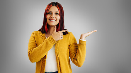 Latin girl with red hair and yellow jacket pointing at something with hands on gray background