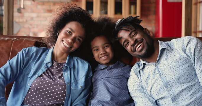 Happy african american couple parents enjoying free tender moment with smiling small mixed race child daughter, relaxing together on comfortable sofa, looking at camera, entertaining indoors.