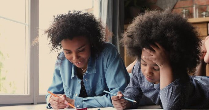 Affectionate loving young african american mother lying on heated floor with small adorable child daughter, communicating chatting discussing life event while drawing pictures in paper album indoors.