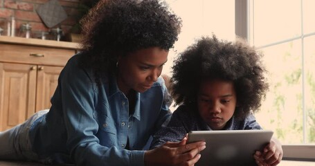Beautiful young african ethnicity woman lying on warm floor with curious small child girl, enjoying playing on digital computer tablet together or involved in educational application usage at home. - Powered by Adobe