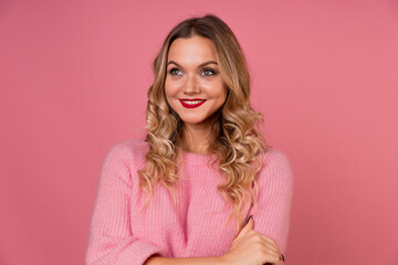 Fototapeta premium Young attractive woman in a warm pink sweater on a pink background, blonde with curly hair and red lipstick. Studio portrait, copy space