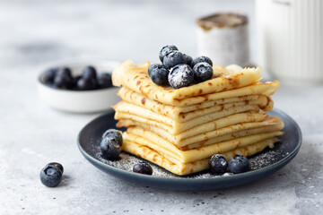 stack of thin pancakes, crepes served with blueberries and powdered sugar on blue plate. Homemade...