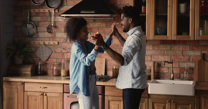 Happy young african woman learning dancing in pairs with affectionate biracial husband, twisting holding hands in kitchen. Joyful positive romantic mixed race family couple having fun indoors.