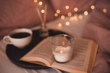 Hygge concept with cup of coffee, open paper book with staying burning candle and glowing lights at background close up. Cozy atmosphere.