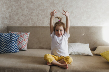 a little girl is sitting on the couch in yellow pants and a white T-shirt and doing morning exercises.