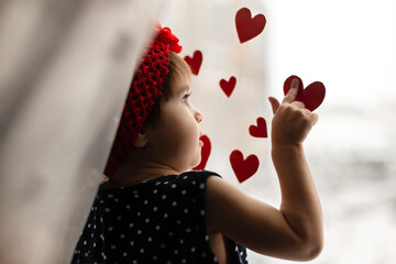 Little girl with small red paper hearts near the window.