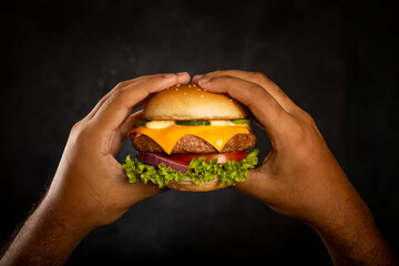 Hands holding cheeseburger on dark background.