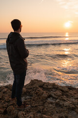 Man looking at the sunset with his sunglasses and the waves crashing in the Atlantic ocean in Portugal