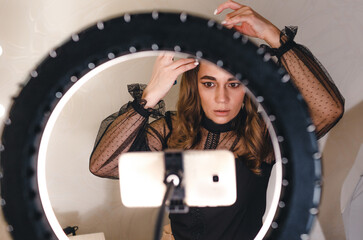 A girl blogger tells how to do her hair to her subscribers looking at the phone, in front of a large lamp.