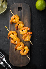 Tasty spiced shrimp skewers  sriracha kebabs with lime, on wooden serving board, on black background, top view flat lay