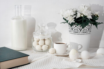 Bottle of milk, book, coconut  candies in a glass jar, cup of espresso coffee, flower pot with chrysanths, eggs, kitchen towel on the table. White wall at the background.  Breakfast still life.