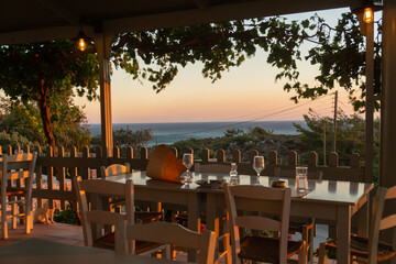 Chairs with tables, empty glass wine in typical Greek tavern in Elafonisi, Crete island, Greece. Plants with hanging fruits in foreground, warm colors of golden hour
