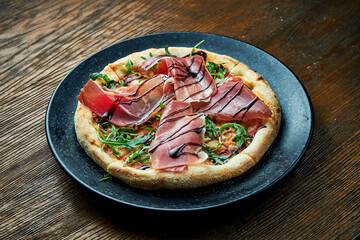 Wood-fired pizza with jamon, arugula, red sauce and balsamic with crispy sides, served on a black plate on a wooden background. pizzette a kind of Italian pizza