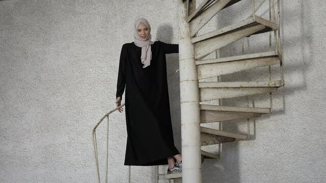 Young woman in long dress and hijab smiles standing on white metal spiral staircase of architectural landmark slow motion