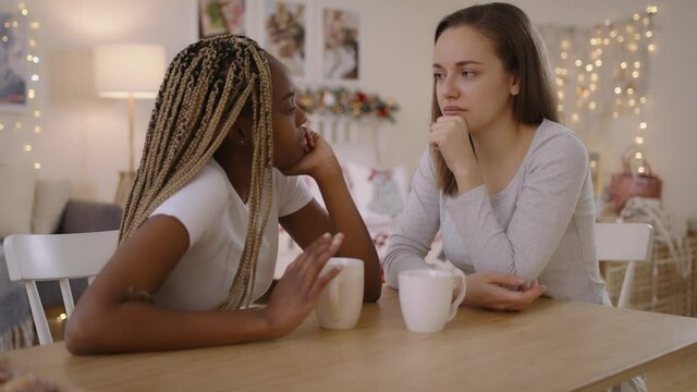Mixed Race Lesbian Couple Portrait. Homosexual lesbian couple chatting sitting at table at home, touching each other, laughing and hugging. Multi-ethnic family spend time together