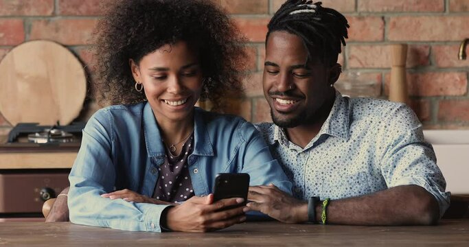 Smiling sincere young african american family couple sitting at table, using mobile software applications or scrolling news feed in social networks, mixed race technology addicted people concept.