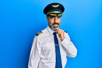 Middle age man with beard and grey hair wearing airplane pilot uniform looking confident at the camera with smile with crossed arms and hand raised on chin. thinking positive.