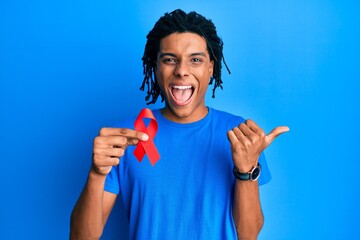 Young african american man holding support red ribbon pointing thumb up to the side smiling happy...